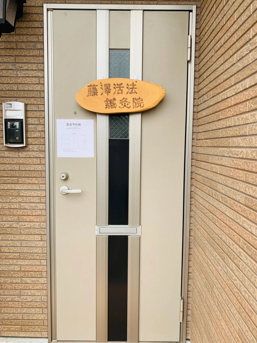 建物の玄関。ドアの上に藤澤活法鍼灸院の木の看板がかかっている